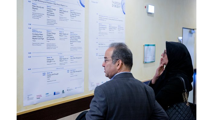 Ein Mann und eine Frau lesen das Konferenzprogramm, das als Poster an die Wand gehängt ist.