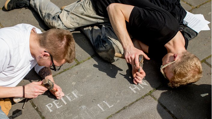 Zwei Personen meißeln Namen in Bodenplatten.