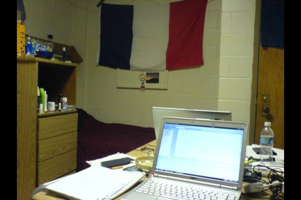 In einem Wohnheimzimmer steht ein aufgeklapptes Notebook auf einem Schreibtisch - im Hintergrund, Kommode, Bett und eine Frankreichflagge an der Wand
