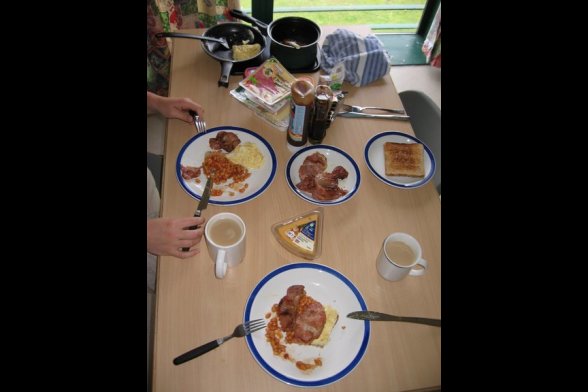 Blick auf einen Frühstückstisch mit Teetassen Toast, Speck und Rührei, Gebackenen Bohnen und Cheddar