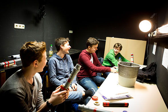Vier Jungen sitzen in einem schwarzen Raum an einem Tisch und lachen.