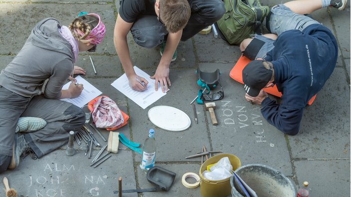 Drei Personen meißeln Namen in Bodenplatten.