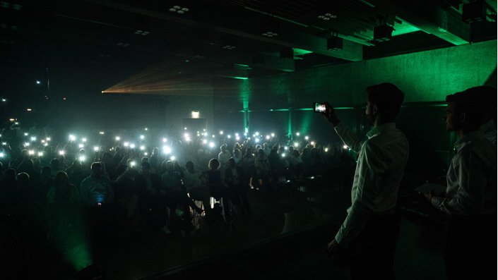 Blick von der Bühne auf den abgedunkelten Zuschauerrraum, in dem zahlreiche Zuschauer ihre Handys leuchten lassen.