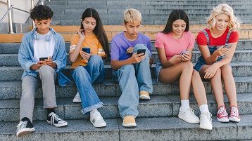 fünf Teenager, die nebeneinander auf einer Treppe sitzen und auf ihre Handys schauen (Bild: Maria Vitkovska / AdobeStock)