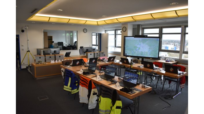 Simulations-Stabsraum des Deutschen Zentrums für Luft- und Raumfahrt (DLR). Auf den Tischen stehen Laptops. Im Hintergrund ist ein großes Display zu sehen, auf dem eine Lagekarte eingeblendet ist. 