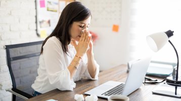 Frau, die am Schreibtisch vor offenem Laptop sitzt und sich die Nase putzt (Bild: AntonioDiaz / Adobe Stock)