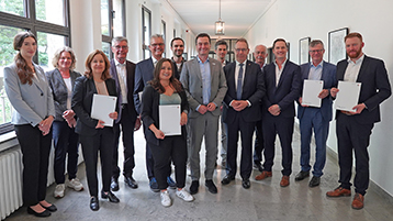14 Vertreter*innen der TH Köln, des Innovation Hub Bergisches RheinLand und der Bezirksregierung Köln stehen in einem weißen Flur und halten die Förderbescheide in die Kamera. (Bild: Bezirksregierung Köln)