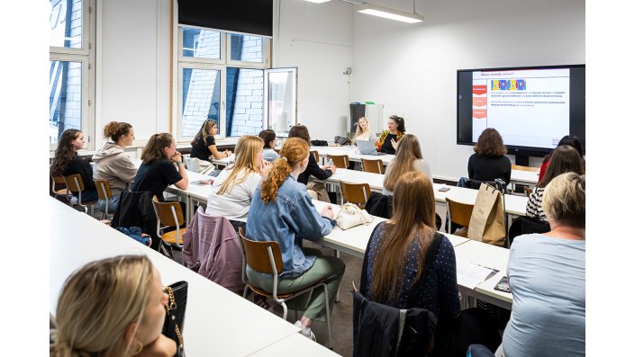 Seminarraum mit vielen Interessentinnen. Am Tisch vorne sitzt eine Professorin und spricht zu den Besucher*innen..