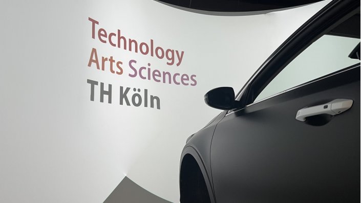 Fahrsimulator vor Leinwand mit Logo der TH Köln