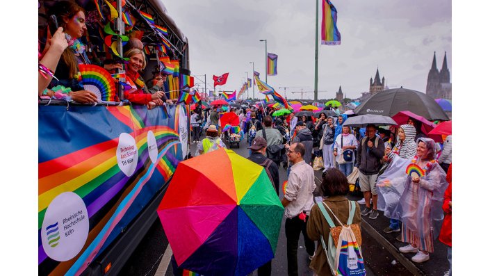 Die CSD-Parade auf der einer Brücke in Köln. Links der Wagen der Hochschulen, rechts Zuschauer*innen.