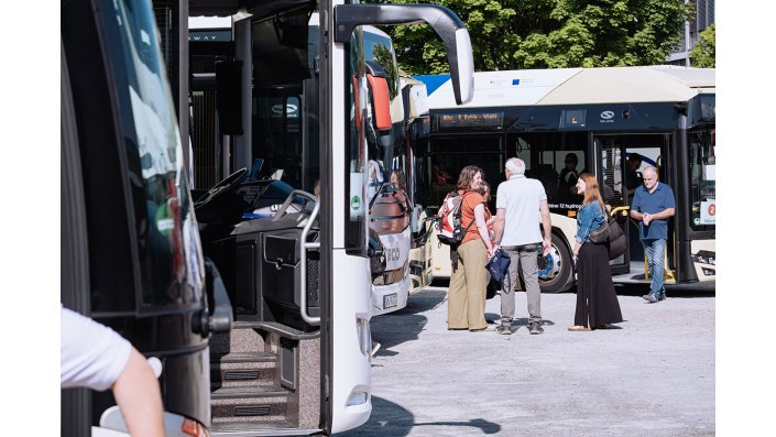 Busse stehen auf einem Parkplatz, auf dem sich ebenfalls fünf Personen befinden, die sich unterhalten.
