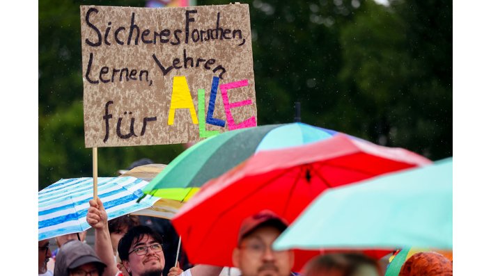 Eine Plakat wird hochgehalten, auf dem steht "Sicheres Forschen, Lernen, Lehren für ALLE"