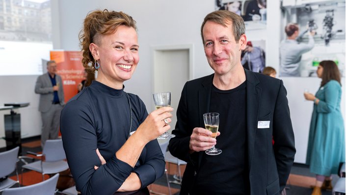 [Foto] Zwei Personen mit Sektgläsern in der Hand lächeln in die Kamera.