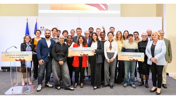 [Foto] Gruppenfoto von 26 Personen auf einer Bühne bei der Verleihung des European Hackathon 2026. Einige halten große Preisschilder: 1. Preis und 3. Preis. Im Hintergrund sind zwei Flaggen und ein Schild mit dem Text „Ministère des Transports“. Ein Rednerpult mit dem Eventnamen steht links.