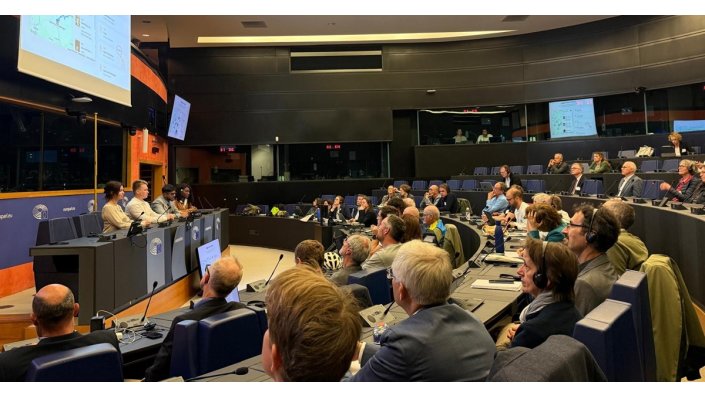 [Foto] Sitzung in einem modernen, halbkreisförmigen Saal mit mehreren Personen auf Stühlen, die einem Podium mit fünf redenden Personen zugewandt sind. Im Hintergrund sind zwei Projektionsflächen mit Präsentationsfolien sichtbar.