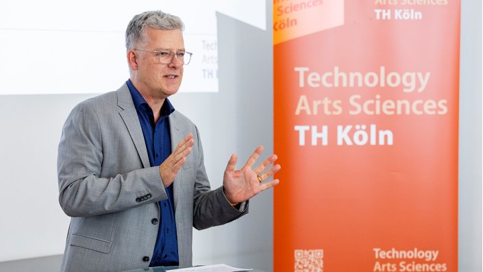 [Foto] Ein Mann mit grauem Jackett steht vor einem Roll-up und gestikuliert beim Sprechen.