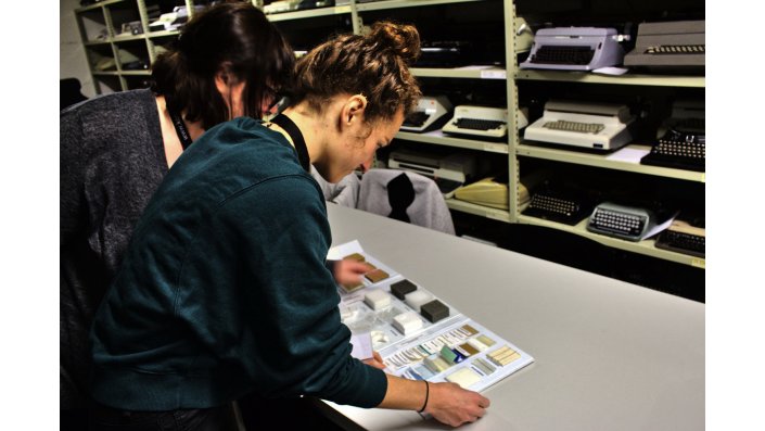 Zwei Studentinnen suchen anhand eines Musterbuchs geeignete Materialien aus.