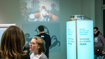 Eine Frau steht vor einer Wand, an der ein Film mit einer Spielzeug-Figur gezeigt wird. Daneben ist eine Leuchtsäule mit dem Schriftzug Institut für Informationswissenschaft Eine Frau steht vor einer Wand, an der ein Film mit einer Spielzeug-Figur gezeigt wird. Daneben ist eine Leuchtsäule mit dem Schriftzug Institut für Informationswissenschaft