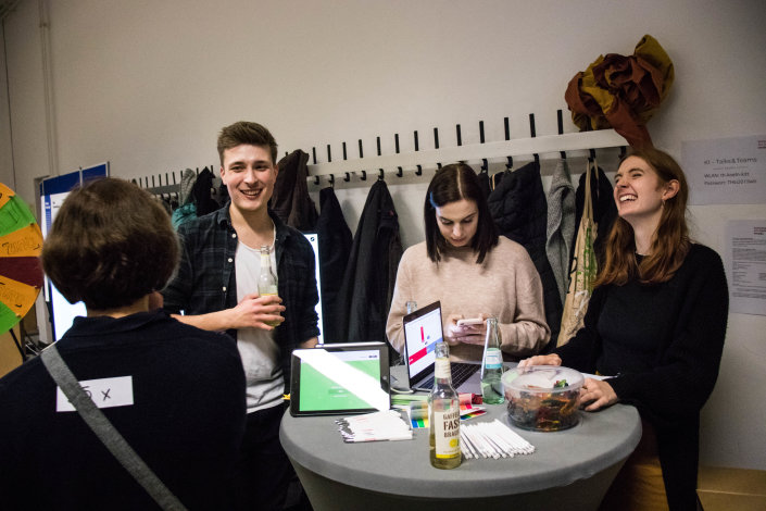 Drei Studierende stehen um einen Stehtisch und lachen, im Hintergrund ist ein Glücksrad Drei Studierende stehen um einen Stehtisch und lachen, im Hintergrund ist ein Glücksrad
