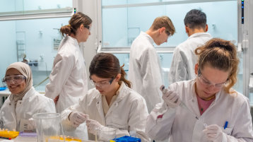 Fünf Nachwuchswissenschaftler*innen arbeiten im Labor und pipettieren, füllen ab und stehen am Abzug. (Bild: TH Köln - Media Lab)