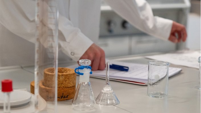 Zu sehen ist eine Werkbank mit Laborgeräten und dahinter die Hand eines Schülers, die einen Kulli hält und auf einen Block stützt.