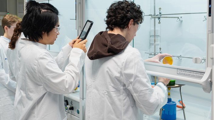 Vier Schülerinnen und Schüler stehen vor einem Abzug im Labor und arbeiten. Ein Jugendlicher fotografiert mit einem Tablet.