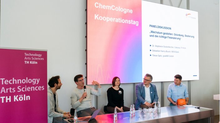 Die fünfköpfige Panelrunde sitzt vor den gefüllten Reihen des Hörsaals 2.