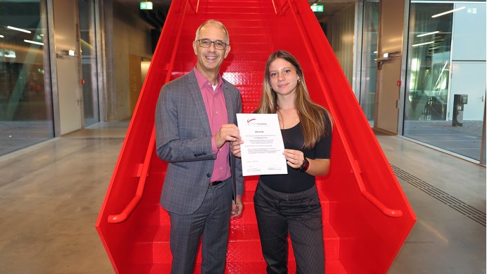 Prof. Dr. Dirk Burdinski und Magdalena Kroll stehen vor der roten Treppe im EG des Campus Leverkusen.