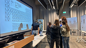 Studierende stehen am Hörsaalpult und schauen auf ein Periodensystem an der Wand (Bild: TH Köln - Media Lab)