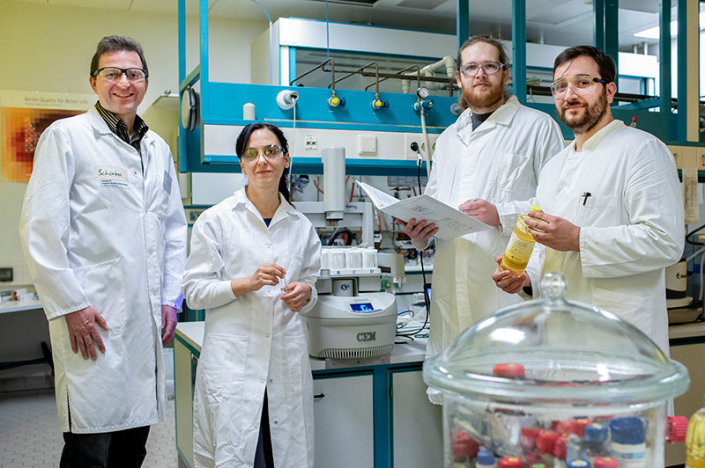 Prof. Dr. Ulrich Schörken, Devrim Eren Seitz, Lars Nitzsche und Tristan Jolmes stehen, mit weißen Laborkitteln bekleidet, in einem Labor