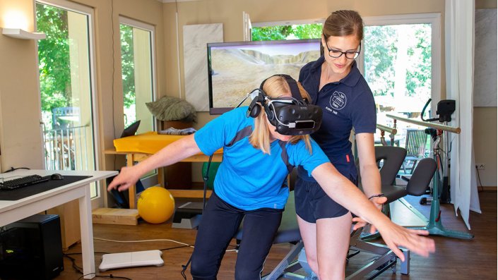 Eine Person mit VR-Brille macht eine Gymnastikübung. Eine weitere Person unterstützt sie dabei.