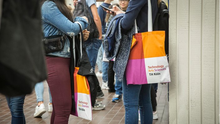 Junge Menschen stehen mit Taschen im Look der TH Köln im Foyer der Hochschule