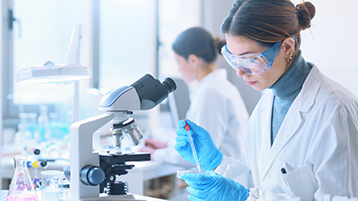 Eine Laborantin mit weißem Kittel und Schutzbrille befüllt eine Petrischale mit einer Pipette und sitzt dabei vor einem Mikroskop. (Bild: Adobe Stock)