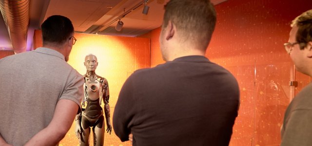 [Foto] Drei Männer von hinten betrachten einen humanoiden Roboter mit metallischem Körper in einem orange beleuchteten Raum. (Bild:Deutsches Museum/Eric Lichtenscheidt)