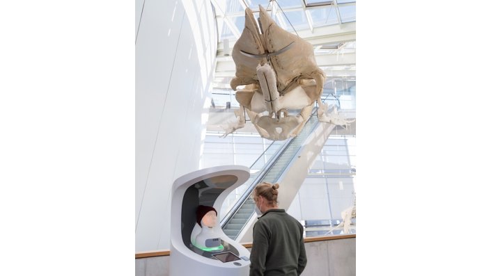 [Foto] Mann steht vor einem interaktiven Kiosk mit Screen. Über ihm hängt ein großes Skelett eines Wals, das von der Decke eines modernen Gebäudes heruntergelassen ist. Im Hintergrund ist eine Glasdecke und eine Rolltreppe zu erkennen.