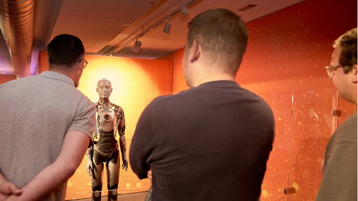[Foto] Drei Männer von hinten betrachten einen humanoiden Roboter mit metallischem Körper in einem orange beleuchteten Raum.