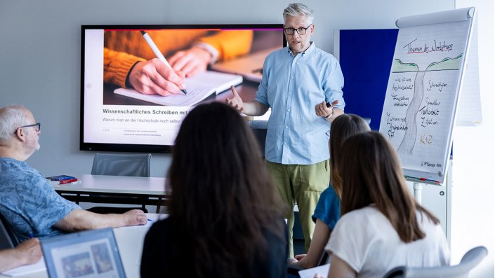 [Foto] Ein Mann mit grauen Haaren und Brille steht vor vier sitzenden Personen. Hinter ihm ist ein Bildschirm, auf dem "Wissenschaftliches Schreiben" steht.