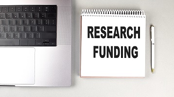 Notizblock, auf dem "Research Funding" steht, neben einem Stift und einem Laptop (Bild: AdobeStock / Iryna)