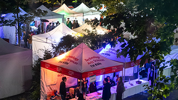 Foto: Mehrere kleine Verkaufszelte und Infostände bei Nacht unter Bäumen im Freien, beleuchtet mit bunten Lichtern, mit mehreren Personen in und um die Zelte. (Bild: Heike Fischer / TH Köln)