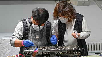 Mr. Yang Seog Joong und Mrs. Lee Yun Jeong bei der Arbeit. Die Aufgaben bestanden in der Beseitigung eines sekundären und objektgefährdenden Überzugs sowie in der Stabilisierung der mit Perlmutteinlagen dekorierten Lackarbeit. (Bild: TH Köln - CICS - Andreas Krupa)