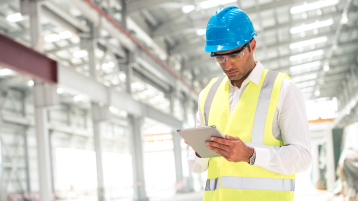 Construction foreman on construction site, holding digital tablet. He is wearing reflective vest over a white shirt, protective helmet and protective glasses. He is checking construction site. Copy space. (Bild: iStock.com/Azmanl)