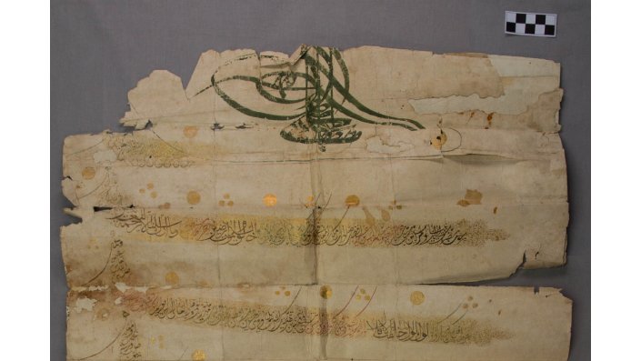 Bild 1 zeigt den oberen Teil des Schriftstückes auf grauem Hintergrund. Zu sehen ist die aufwändig ge-schlungene Tughra des Sultans, der Anfang des Textes samt den goldglänzenden Verzierungen und Schäden wie Fehlstellen, Risse und alte Papierergänzungen.