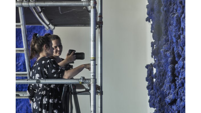 Zwei Studierende, links im Bild, stehen auf einem Gerüst und richten eine Fotokamera auf ein blaues Schwammrelief von Yves Klein, rechts im Bild. Die vordere Studierende zeigt mit der Hand in Richtung des Reliefs und scheint der hinteren etwas mitzuteilen, diese lächelt und hält ihren Blick auf die Kamera gesenkt.