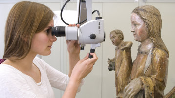 Eine Studierende untersucht mit dem Stereomikroskop eine Skulptur der Heiligen Maria mit dem Kind. (Bild: TH Köln - CICS)