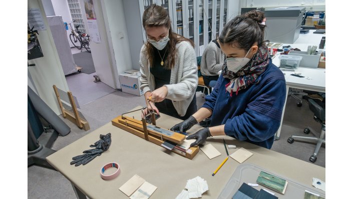 Im Bild wird mit einem Crockmeter die Reibechtheit der gefärbten Täfelchen überprüft. Dabei wird in einem definierten und mit definiertem Druck ein mit einem hellen Textilflicken bestückter Stempel über die Oberfläche des Täfelchens gerieben. Auf dem Textil sammelt sich der Abrieb und auf dem Brettchen lässt sich die Reibespur betrachten.