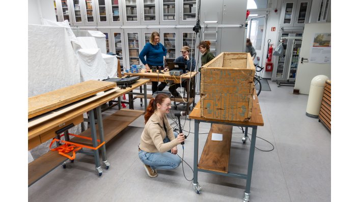 Zur Ermittlung der Eigenfrequenz des Sarges schlägt Corinna Mally mit dem Modalhammer an die Tischkante. Im Hintergrund werden die Resonanzen am Computer von Jakob Becker erfasst. Dr. Kerstin Kracht und Anna-Lena Korsten schauen zu.