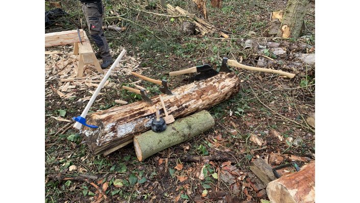 Stillleben im Wald. Hier sind einige der schweren Handwerkzeuge zu sehen: links ein Dechsel mit langem Stiel, in der Mitte mehrere, unterschiedliche Breitbeile und rechts eine langstielige Axt.