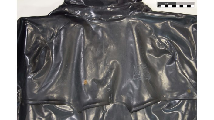 Detail des Schultersattels des schwarzen PVC-Mantels vor der Reinigung mit weißlichen Rückständen von Löschwasser und Rostflecken. (Inventarnummer: K-2015-00022).