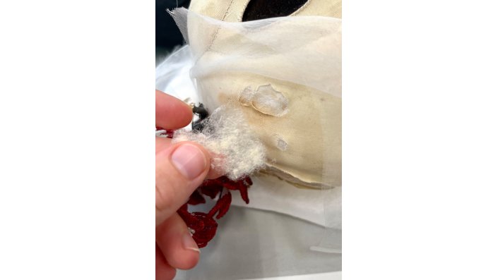 Eine Hand hält ein Stück crèmeweißes Seidenvlies. Im Hintergrund sind Fehlstellen in der "Seidenhaut" an "Desdemonas" Kopf zu sehen.