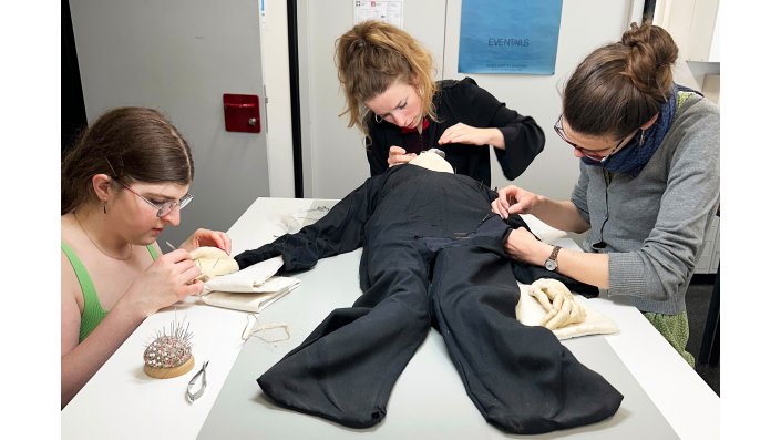 Drei Restauratorinnen arbeiten an der textilen Figur "Desdemona". Die Figur liegt auf dem Tisch und wird simultan am Kopf, an den Händen und am Körper restauriert.